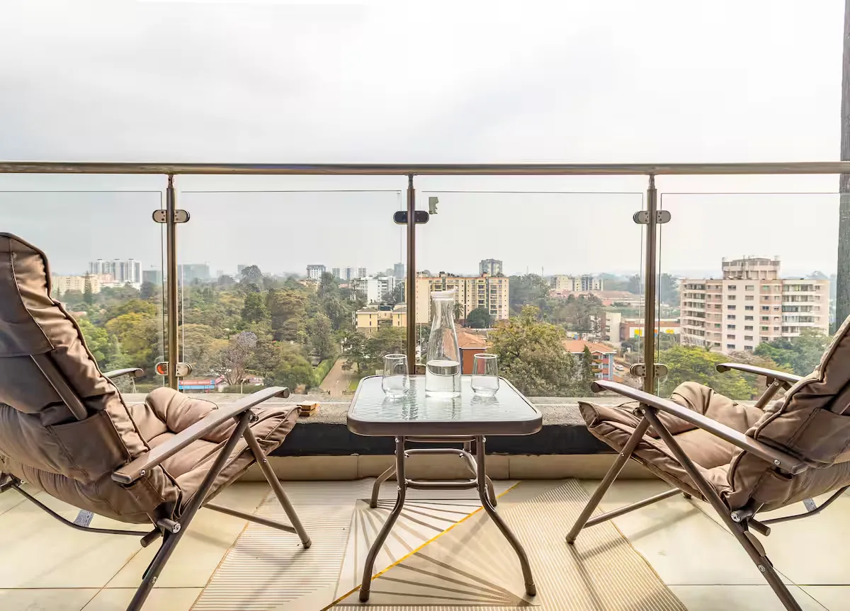 Two reclining chairs and a glass table on a balcony overlooking trees and nearby buildings